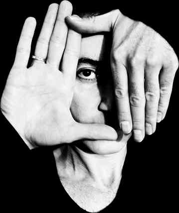 Black and white headshot photograph of a man with his hands obscuring part of his face, making his eye visible.